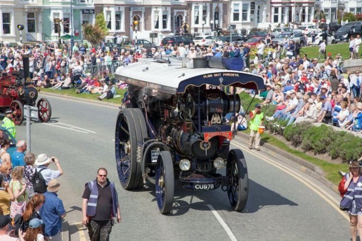Vintage steam tractor in parade surrounded by crowd on sunny day, with rows of houses in the background.