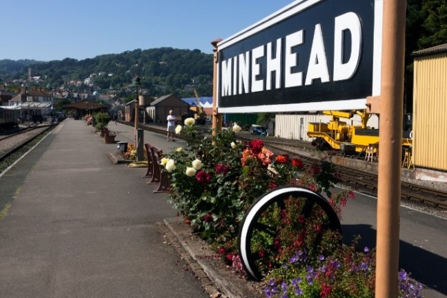 Minehead train station platform with flowers and a sign, under a clear blue sky.