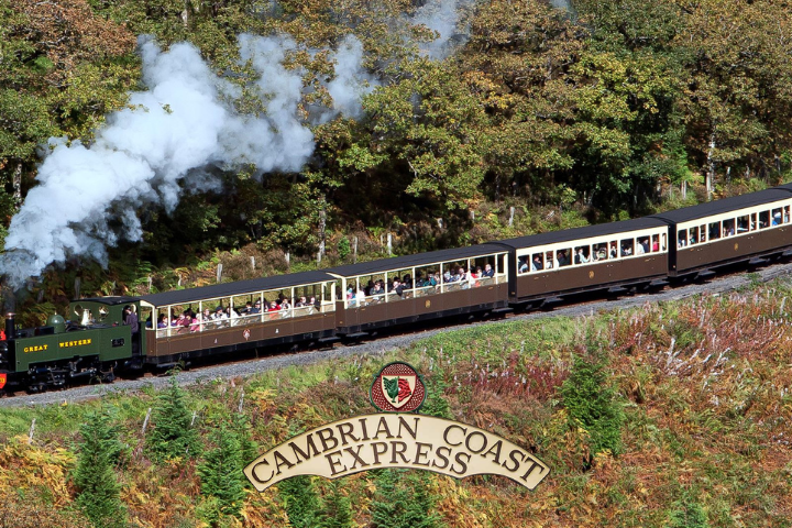 Steam train on a curved track through forested area, sign reads 'Cambrian Coast Express'.