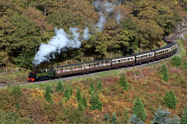 Steam train with passengers in scenic landscape with trees and hills.