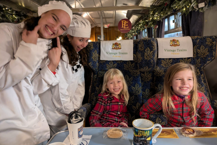 Two children in pajamas and two chefs on a decorated train, with mugs and cookies on the table.