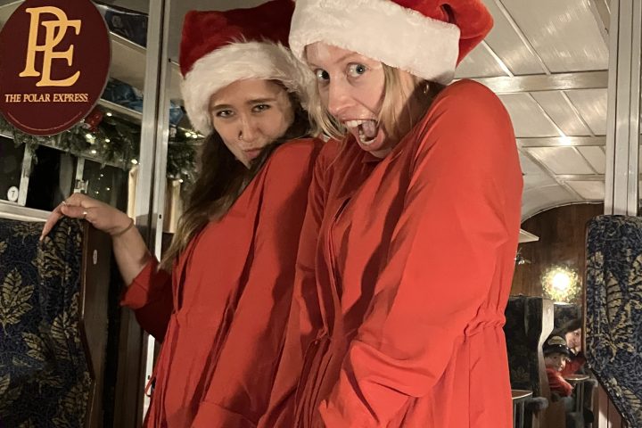 Two people in Santa hats and red outfits posing inside a decorated train with 'Polar Express' sign.