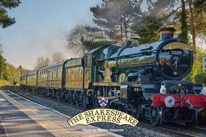 Vintage train 'The Shakespeare Express' at Henley-in-Arden station with steam visible, surrounded by trees.