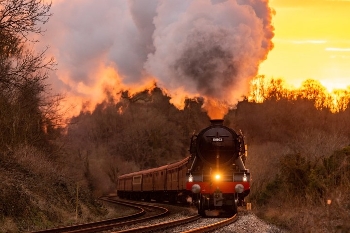 Steam locomotive 60103 emits smoke against a sunrise backdrop, curving through a forested area.