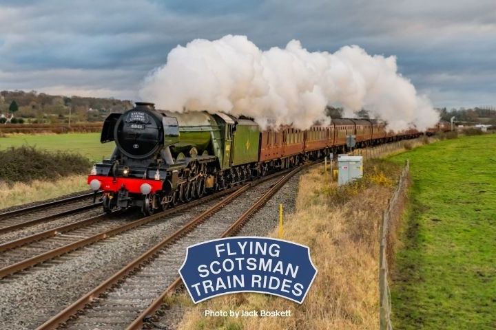Steam train with billowing smoke travels through countryside, grassy fields, and trees in background.