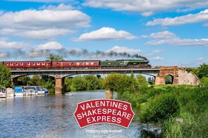 Steam train crossing a bridge over a river with boats, under blue skies with clouds.