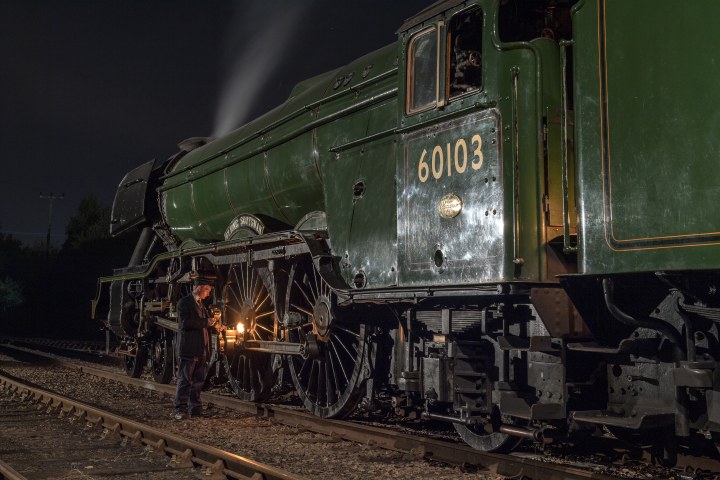 Person in uniform lighting a steam train in a dimly lit train yard at night.