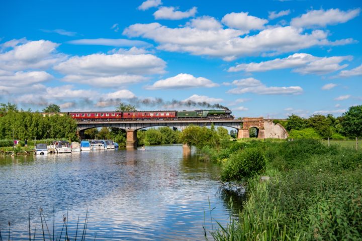 Steam train on bridge over river with boats and green landscape on a sunny day.