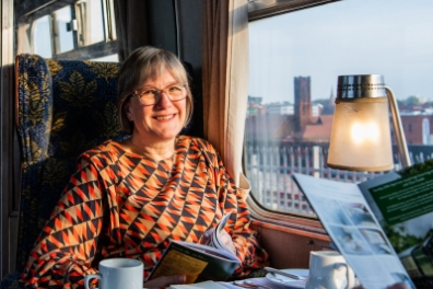 Woman in patterned dress reading on a train with a table set for dining by the window.