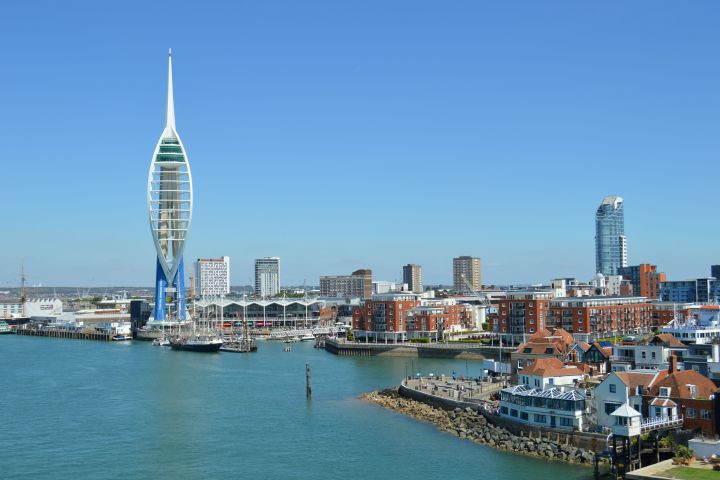 Skyline with a tall spinnaker tower by water, surrounded by buildings under a clear sky.