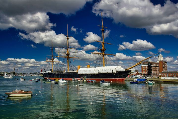 Historic ship docked near a marina with small boats under a partly cloudy blue sky.