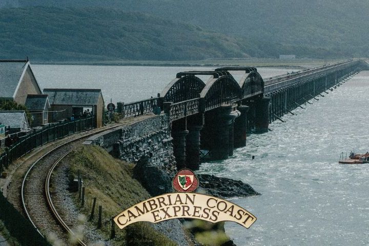 Long railway bridge over water with hills in background and 'Cambrian Coast Express' sign in foreground.