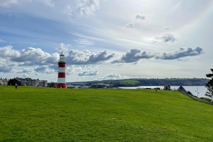 Lighthouse on grassy hill with cloudy sky and distant coastline view.