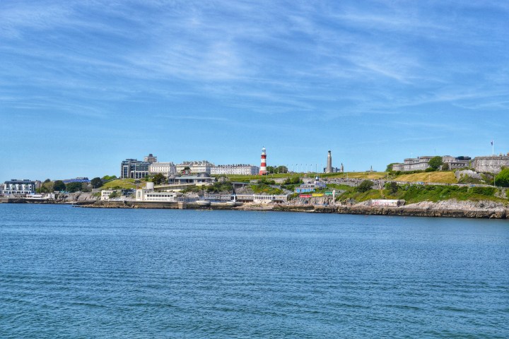 Coastal cityscape with a lighthouse and buildings on a grassy hill under a clear blue sky.