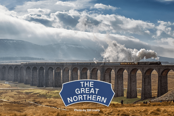 Train crosses large viaduct with mountains in background under cloudy sky.
