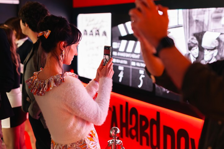 Woman in floral dress and white sweater takes a photo at an exhibit with bright red and black colors.