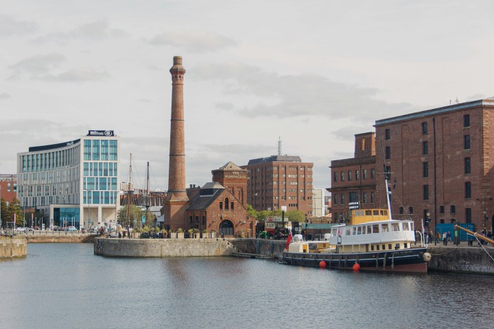 Boats docked by historic buildings and a tall chimney by the water.